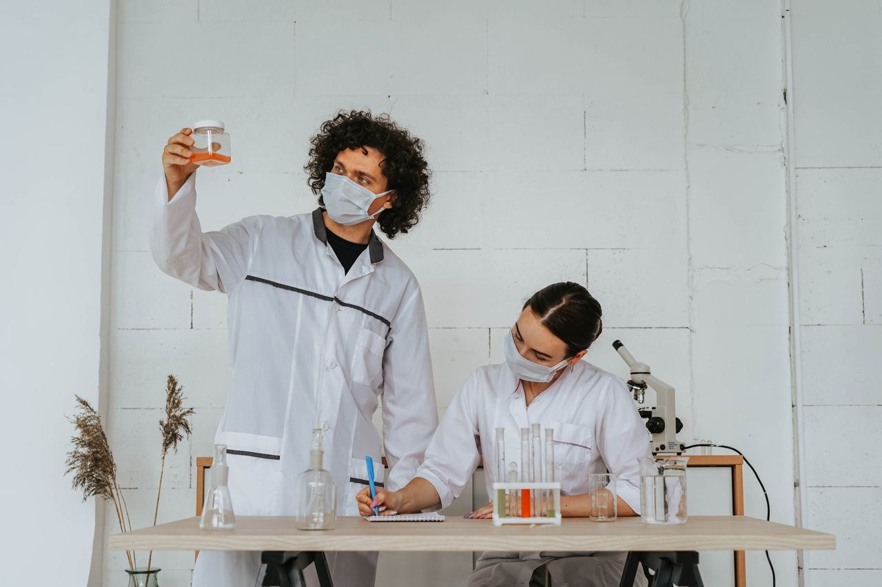 Two scientists in lab coats and face masks conduct an experiment in a laboratory setting.