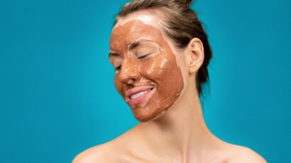 A woman smiles while applying a clay mud mask for skincare treatment.