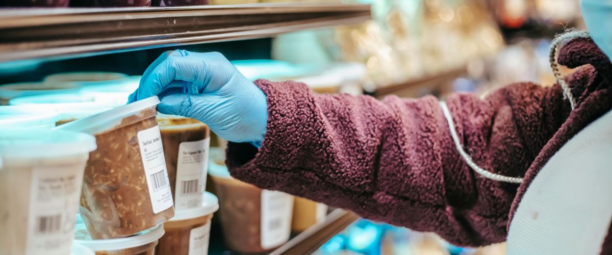 Crop anonymous female customer in protective mask reading label on frozen food in plastic container in grocery store