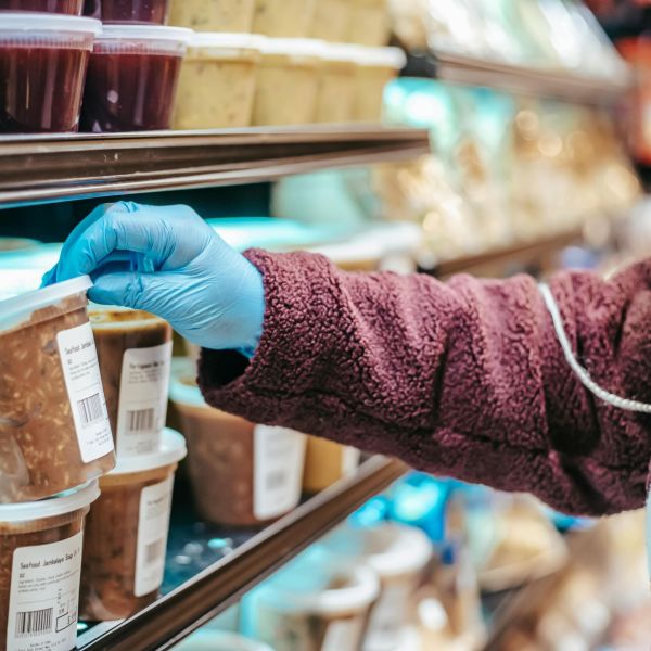 Crop anonymous female customer in protective mask reading label on frozen food in plastic container in grocery store